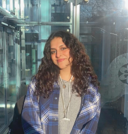 Liba has long curly brown hair and smiles against a glass fronted building